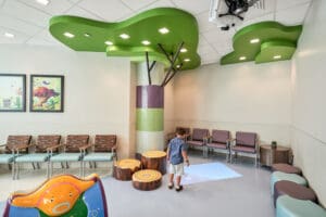 A young child plays in a brightly lit waiting area with tree-themed decorations, including green ceiling panels and stump-shaped seats. This unique playground features colorful seating lining the wall, with framed artwork and a small round table with purple chairs in the background.